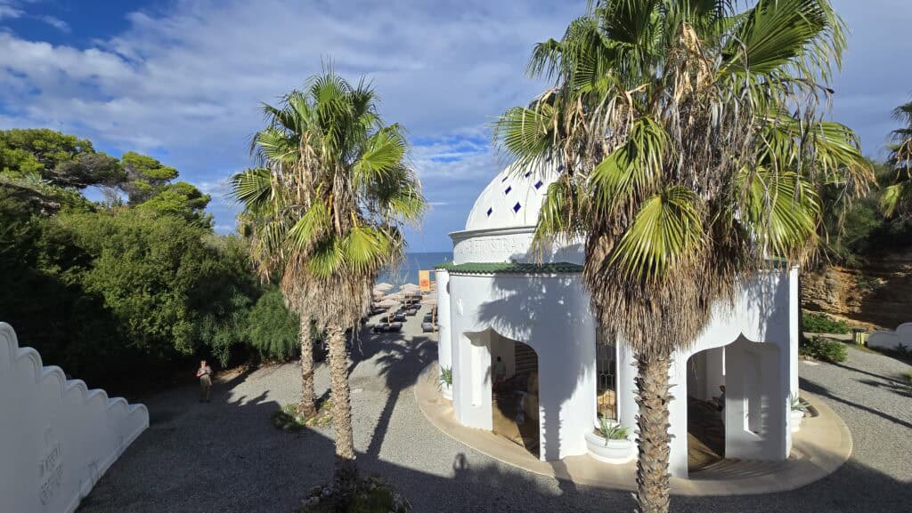 Majesty Reborn: Kallithea Springs Rhodes โ Italian Thermal Baths, Beaches & Architecture 2 Iconic white Rotunda building at Kallithea Springs, Rhodes, framed by palm trees, with sunbeds visible on the pebble beach and the turquoise Aegean Sea in the background.