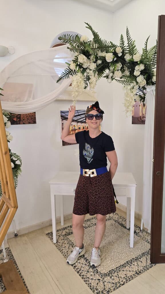 Majesty Reborn: Kallithea Springs Rhodes โ Italian Thermal Baths, Beaches & Architecture 5 Ila posing under a decorative wedding arch made of pink and white flowers and greenery, inside one of the white-walled rooms at Kallithea Springs.