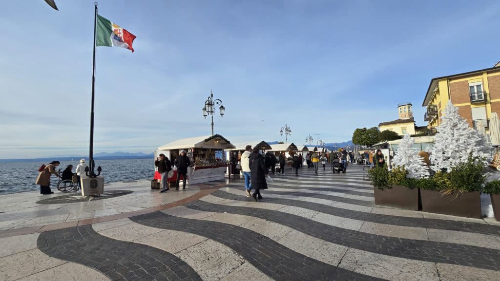 A wide-angle photo showing the Christmas market stalls and crowds lining the Lazise lakeside promenade on Lake Garda. People are walking between the brightly lit stalls, with the railing and the water clearly visible, confirming the market's scenic waterfront location.
