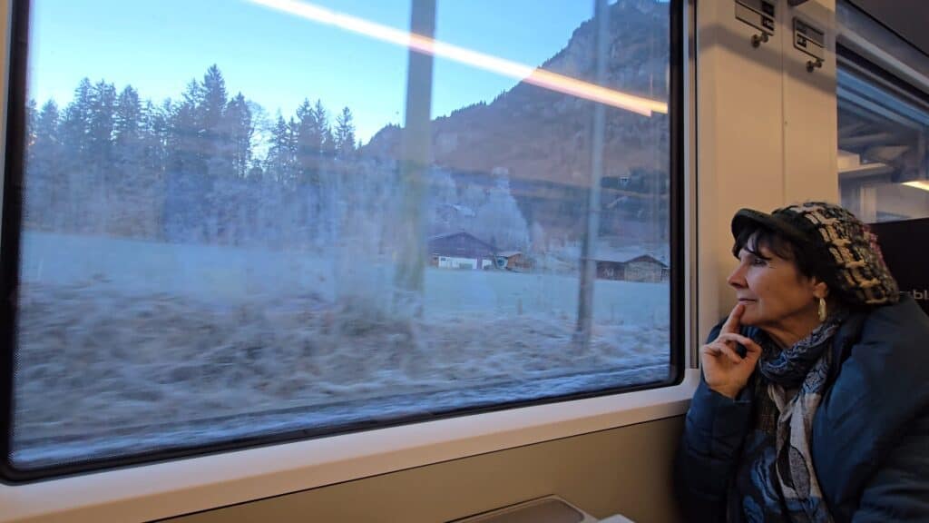 A traveler looking out of the window of the Green Train of the Alps, showing a snowy Alpine landscape and traditional chalets during a winter trip curated by Ila from Tripilare.