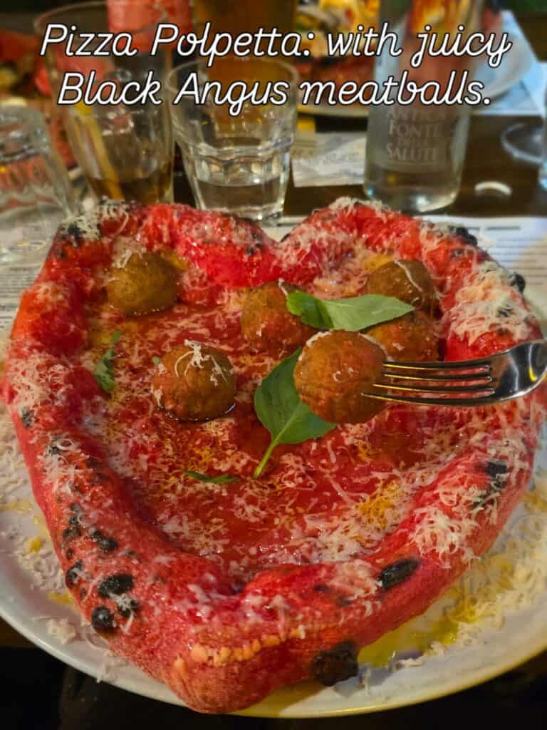 A close-up of a heart-shaped pizza with red dough, topped with Black Angus meatballs, tomato sauce, and grated provola.