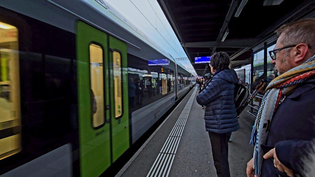 A silver and lime-green BLS train arriving at a platform in Bern railway station, with passengers including Gabriel and author Ila waiting to board.