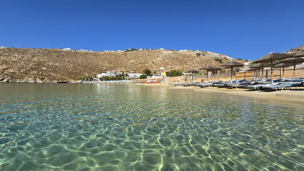 A wide view of Psarou Beach in Mykonos taken from the crystal-clear turquoise water, showing the contrast between the modest Cavo Psarou sunbeds, the small free beach area, and the luxury Nammos umbrellas under a bright blue sky.