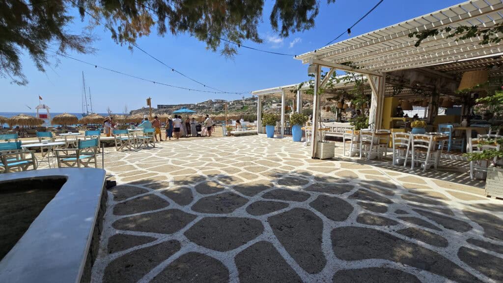 Daytime view of the stone-paved square at Super Paradise Beach in Mykonos, featuring Taverna Talos on the right and the beach in the background.