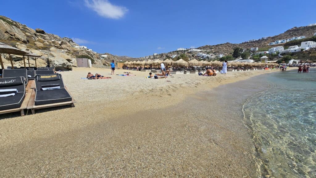 A morning view of the free beach section at Super Paradise Beach, Mykonos, with people laying towels on the sand and Gabriel in blue swimwear standing near the shore.
