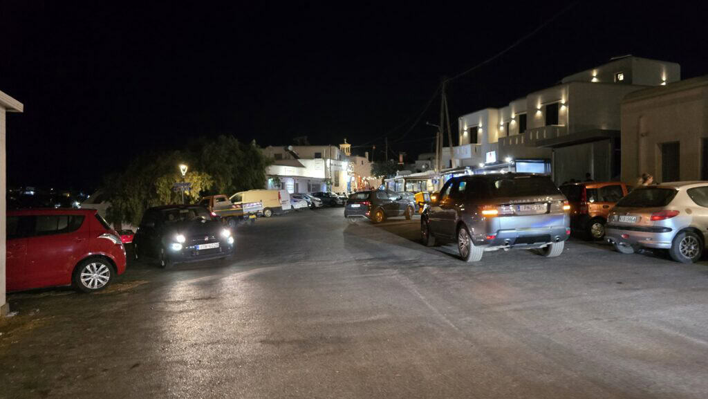 A crowded outdoor parking lot in Ano Mera, Mykonos, at night in August 2025, showing several cars waiting for a spot to open up.