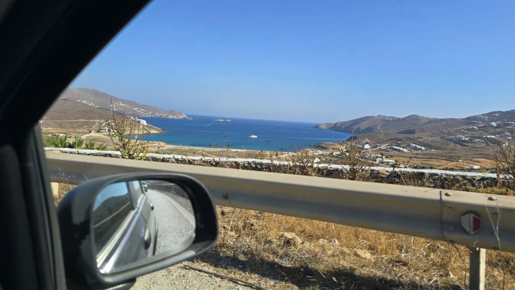 View from inside a rental Fiat Panda driving the scenic main road from Ano Mera to Mykonos Town, showing the blue Aegean sea and the car reflected in the side mirror.