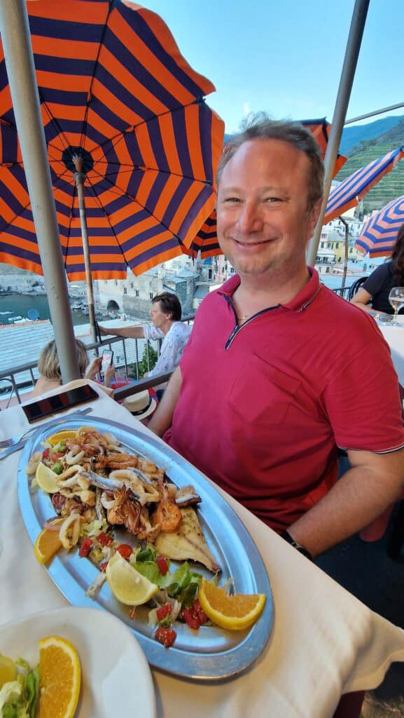 Vernazza Italy: A Gem of the Cinque Terre 8 Gabriel enjoying a fresh seafood dinner at a terrace restaurant in Vernazza with a view of the harbor and colorful umbrellas during sunset.
