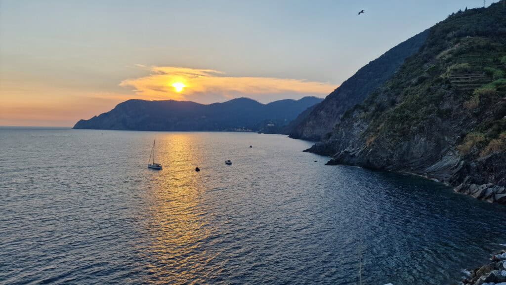Vernazza Italy: A Gem of the Cinque Terre 10 A breathtaking golden sunset over the Mediterranean Sea, viewed from an elevated restaurant terrace in Vernazza, Cinque Terre, with small sailboats anchored in the shimmering water and rugged cliffs in the background.