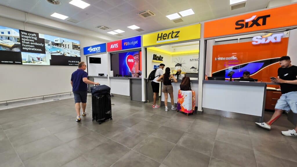 Gabriel waiting with luggage at the Mykonos Airport car rental kiosks, showing the process of picking up a vehicle at arrival.