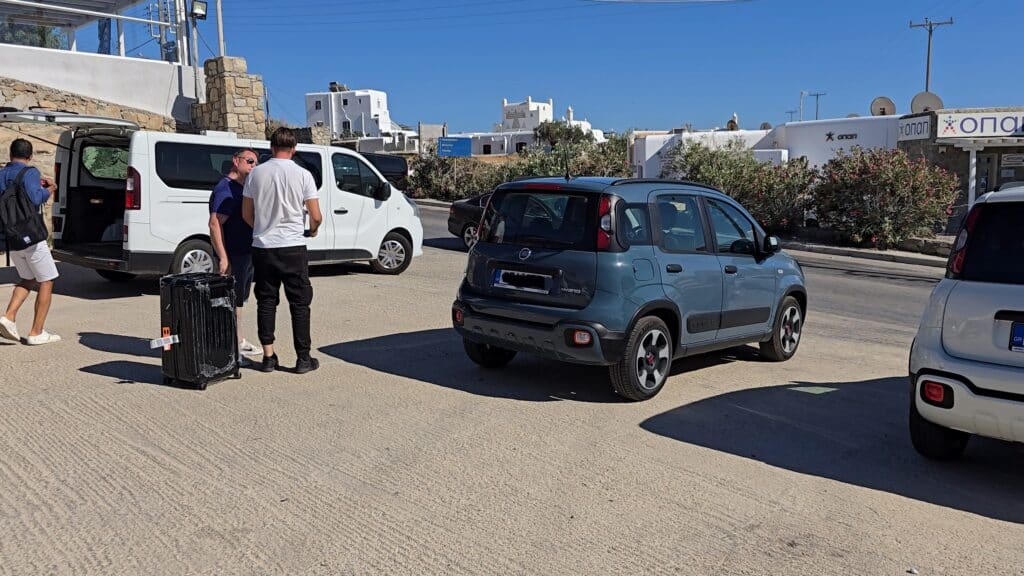 A staff member from the car rental agency explains the vehicle's features to Gabriel, who is standing with a black suitcase next to a blue Fiat Panda in a parking area.
