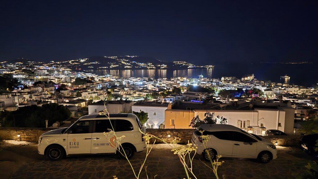 A high-angle night view of Mykonos Town with glowing lights and cars parked along the winding hillside roads leading to the center.