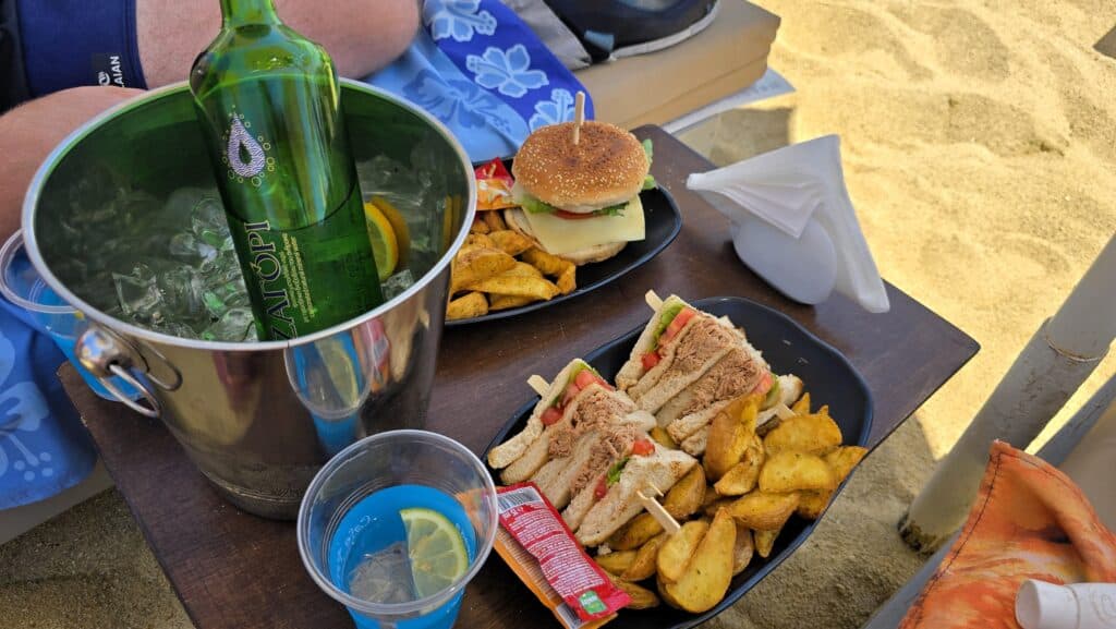 A personal perspective from a sunbed at Paraga Beach, Mykonos. A small wooden beach table sits between two sunbeds, holding a lunch of a hamburger with fries, a club sandwich, and a bottle of water. The golden sand of the beach is visible in the background under a bright blue sky, illustrating the premium beach service of August 2025.