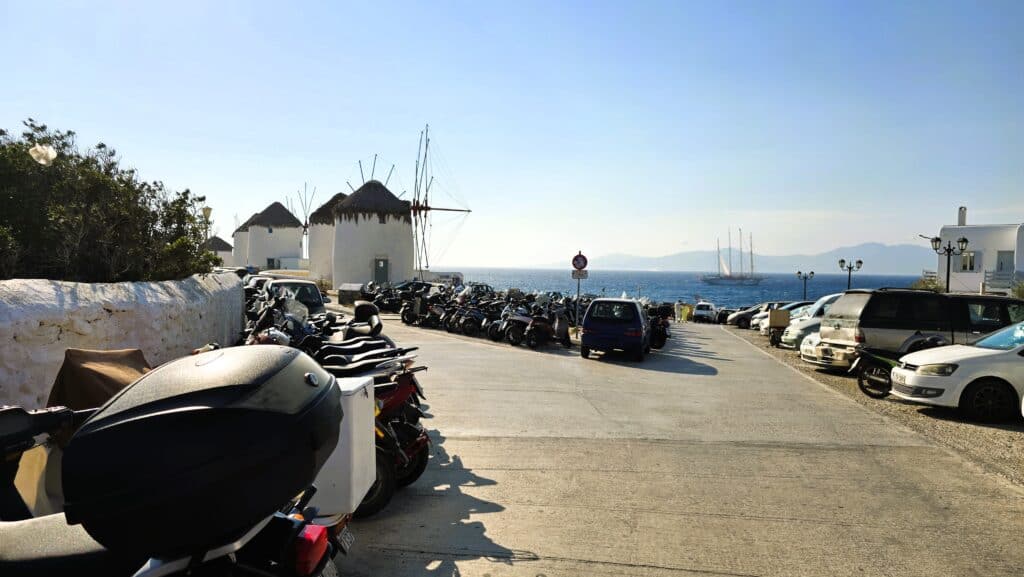 A small white rental car parked in a tight corner among dozens of scooters at Mykonos windmills, used as a visual example of how a compact vehicle can fit into limited spaces during peak season.