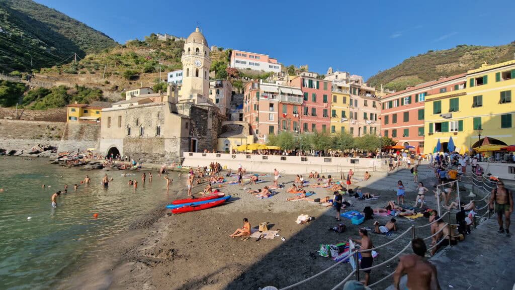 Vernazza Italy: A Gem of the Cinque Terre 4 Crowded small beach in Vernazza harbor at 6:30 PM in August, with people sunbathing and the Santa Margherita di Antiochia Church in the background.