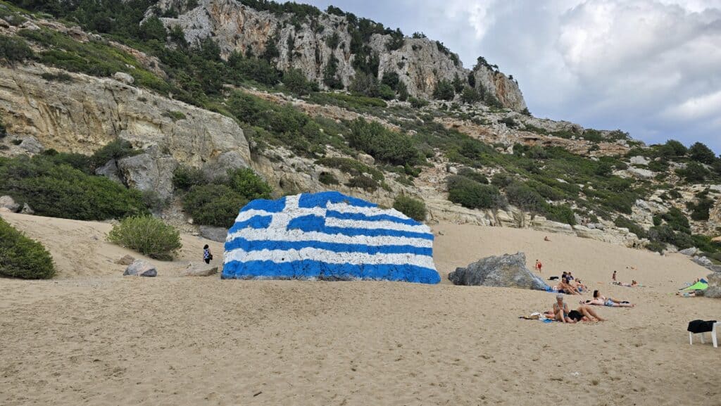 A large Greek flag painted in blue and white stripes across a prominent boulder on the beach at Tsambika, Rhodes, with a steep sandy slope (dune) rising behind it towards the rocky headland.