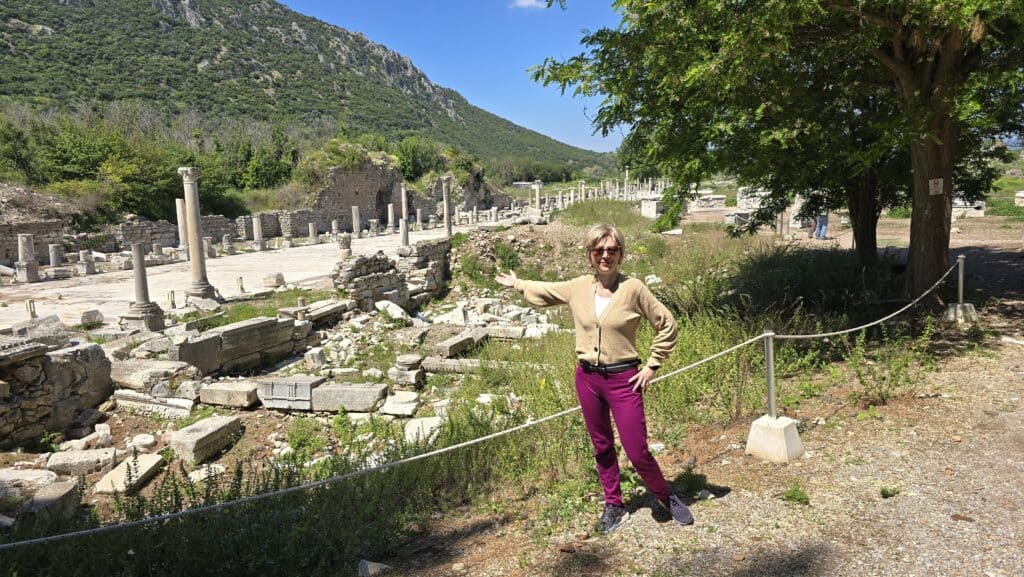 Ephesus & Kusadasi: A Sanctuary of History and the Soul of the Aegean 2 Ila, the author of Tripilare, dressed in a tan cardigan and pink pants, standing and gesturing toward the historic Harbor Road in Ephesus.