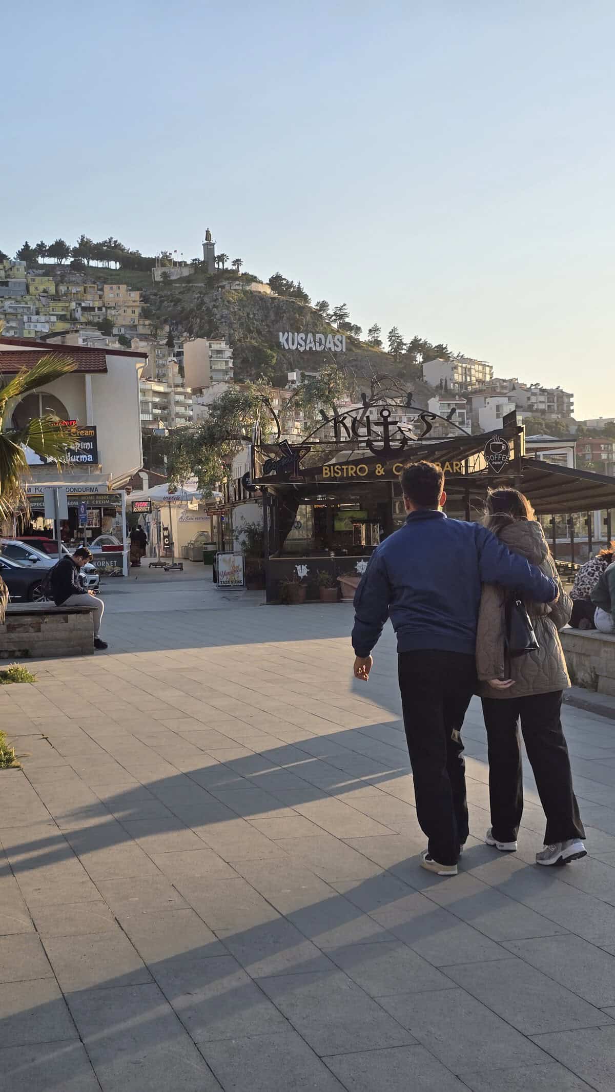 A couple walking embraced along the Kusadasi waterfront promenade with the hillside town and Kusadasi sign in the background.