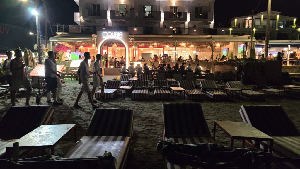 Laganas, Zakynthos: The Honest Truth – Beyond the Tourist Hype 3 People walking and socializing on the sandy beach of Laganas, Zakynthos, in front of a brightly lit beach bar at night.