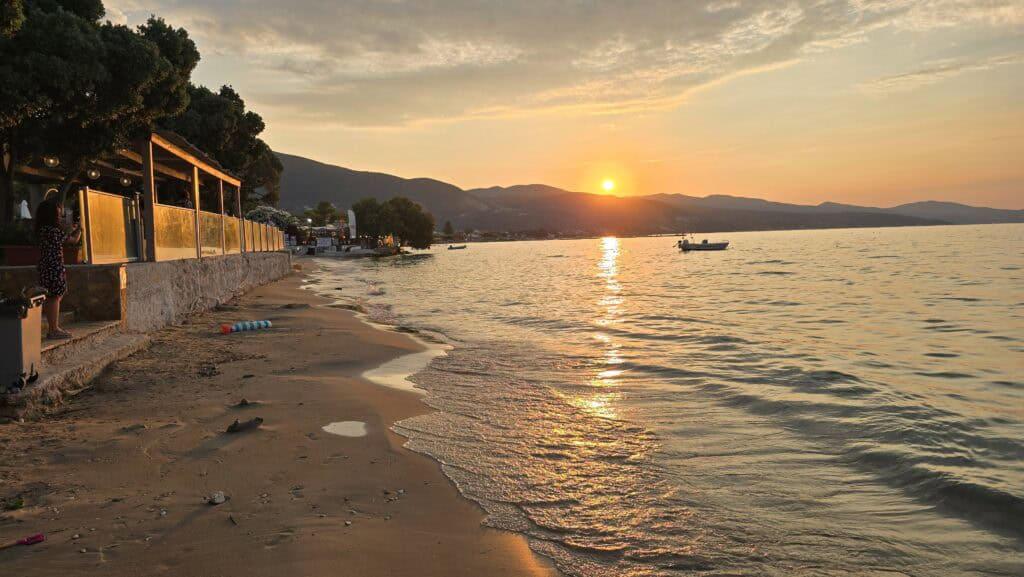 Sunset over calm waters and golden sand at Alykanas Beach in Zakynthos, Greece, with hills and a small boat in the background.