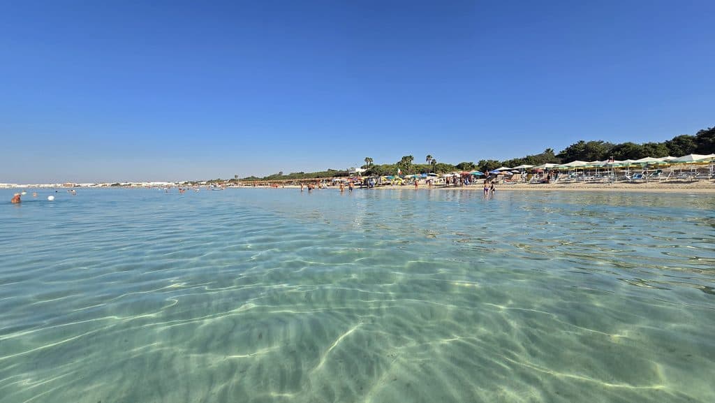Experiencing Torre Lapillo, Salento, Puglia: Honest Travel Guide (Beach, Costs, & Real Experience) 1 Crystal clear shallow water and sandy shoreline at Torre Lapillo beach in Puglia, Italy.